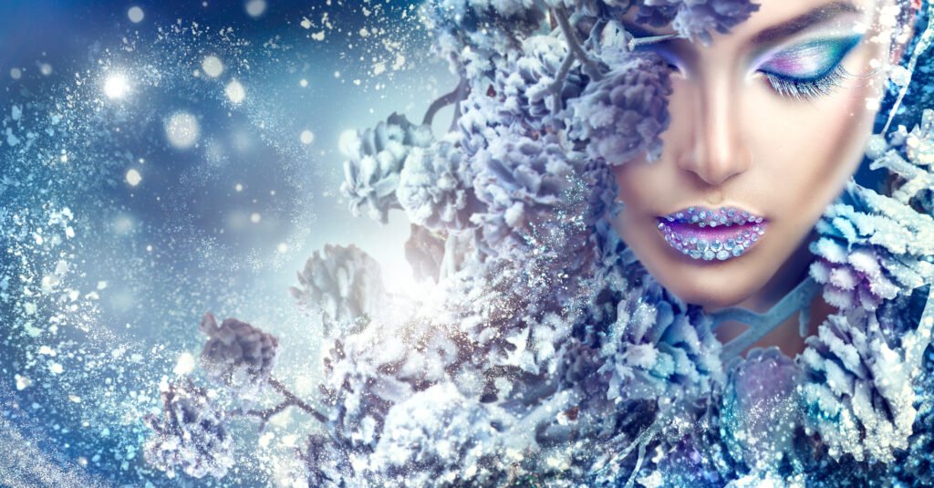 Winter-inspired fashion portrait with a woman wearing icy blue and purple makeup and gemstone-adorned lips surrounded by snow-covered branches.