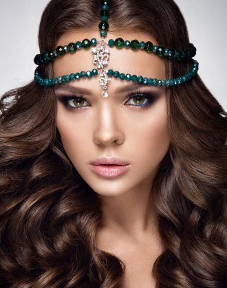 Close-up of a model with long wavy brown hair wearing an emerald and sapphire beaded headpiece with a diamond pendant centerpiece, showcasing a luxurious and mystical look.