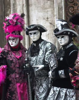 Group of four individuals dressed in intricate Venetian masquerade costumes, featuring elaborate embroidery, feathers, and ornate masks, set in a historic setting with baroque architecture.