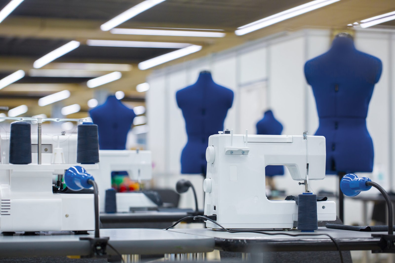 A professional sewing studio with white sewing machines and navy blue dress forms arranged in the background, emphasizing a clean and organized workspace for garment production and design.