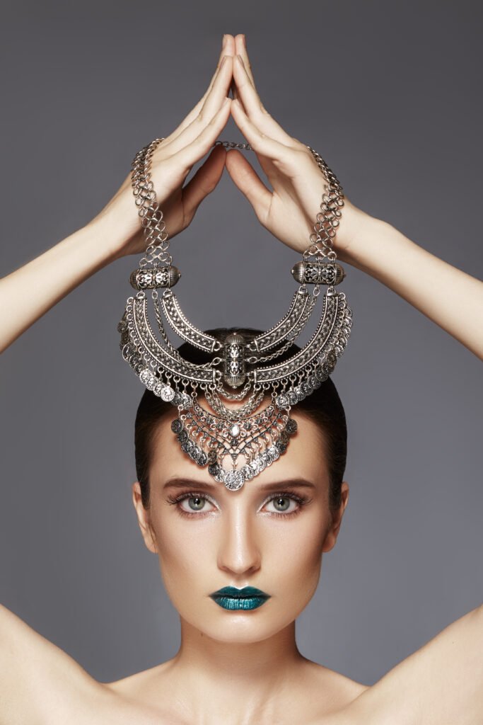 Model holding an intricate silver statement necklace above her head, featuring bold geometric designs and detailed metalwork, paired with striking teal lipstick.