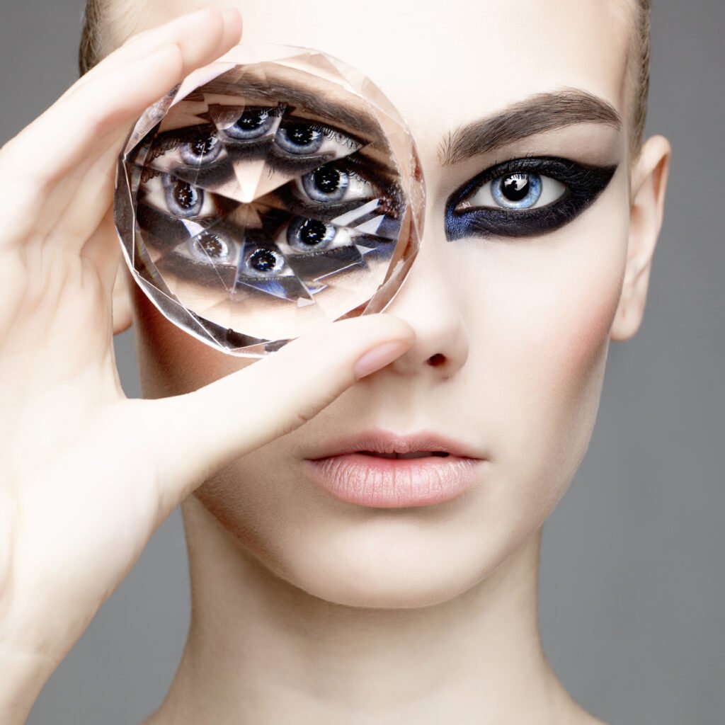 Model with bold black eye makeup holding a large faceted crystal in front of one eye, creating a kaleidoscope effect of multiple eye reflections for a surreal, artistic look.